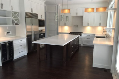 This is an example of a large contemporary u-shaped kitchen in Vancouver with dark hardwood flooring, a belfast sink, shaker cabinets, white cabinets, composite countertops, white splashback, glass sheet splashback, stainless steel appliances and an island.