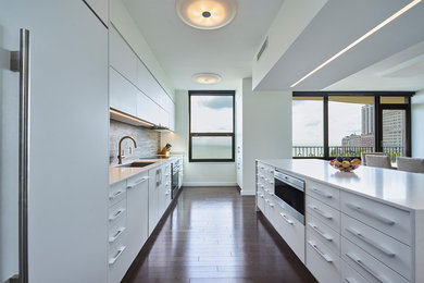This is an example of a large modern single-wall kitchen/diner in Chicago with a single-bowl sink, flat-panel cabinets, white cabinets, engineered stone countertops, grey splashback, porcelain splashback, integrated appliances, dark hardwood flooring, an island, brown floors and white worktops.