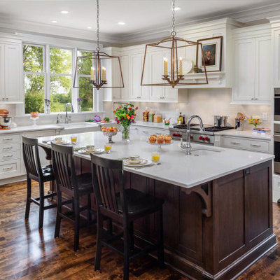 Elegant l-shaped dark wood floor and brown floor kitchen photo in Milwaukee with a farmhouse sink, shaker cabinets, white cabinets, gray backsplash, subway tile backsplash, stainless steel appliances, an island and white countertops