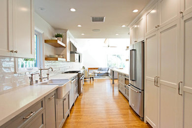 Example of a country galley medium tone wood floor open concept kitchen design in Orange County with white backsplash, stainless steel appliances, no island, white cabinets, a farmhouse sink, shaker cabinets and white countertops