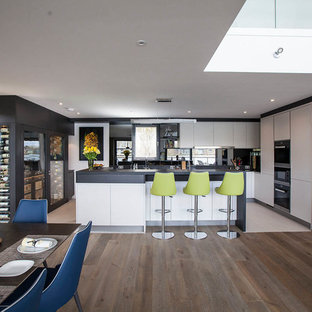 This is an example of an expansive contemporary u-shaped open plan kitchen in Essex with flat-panel cabinets, white cabinets, an island, black worktops, black appliances and white floors.