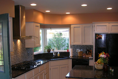 Inredning av ett klassiskt mellanstort kök, med en undermonterad diskho, luckor med infälld panel, vita skåp, granitbänkskiva, blått stänkskydd, glaspanel som stänkskydd, svarta vitvaror och en köksö