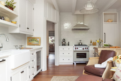 Modelo de cocina clásica con fregadero sobremueble, armarios estilo shaker, puertas de armario blancas, salpicadero blanco, electrodomésticos de acero inoxidable y suelo de madera en tonos medios