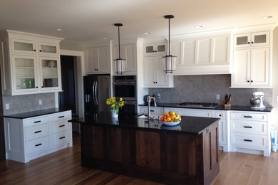 Example of a small trendy medium tone wood floor and brown floor kitchen design in Other with a farmhouse sink, shaker cabinets, white cabinets, gray backsplash, stainless steel appliances and an island