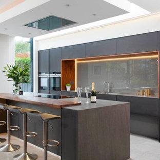Photo of a large modern single-wall separate kitchen in DC Metro with a drop-in sink, flat-panel cabinets, black cabinets, wood benchtops, grey splashback, black appliances, porcelain floors, with island, beige floor, glass sheet splashback and brown benchtop.