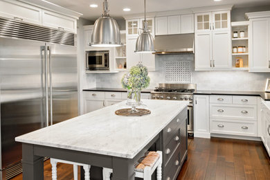 Inspiration for a transitional l-shaped dark wood floor and brown floor enclosed kitchen remodel in New York with a farmhouse sink, shaker cabinets, white cabinets, quartzite countertops, gray backsplash, stone tile backsplash, stainless steel appliances, an island and white countertops