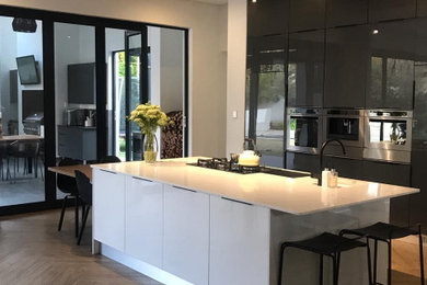 This is an example of a large contemporary l-shaped kitchen with flat-panel cabinets, black cabinets, composite countertops, integrated appliances, vinyl flooring, an island, brown floors and white worktops.