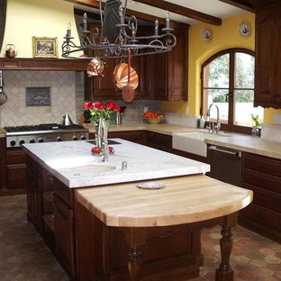 Inspiration for a mid-sized mediterranean l-shaped eat-in kitchen in Los Angeles with a farmhouse sink, raised-panel cabinets, dark wood cabinets, limestone benchtops, grey splashback, brick splashback, stainless steel appliances, terra-cotta floors and with island.