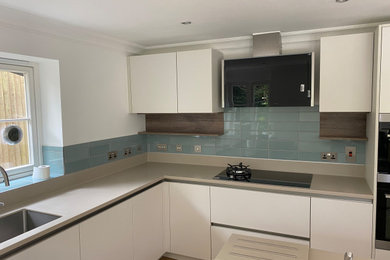 Photo of a large contemporary l-shaped kitchen/diner in Oxfordshire with a single-bowl sink, flat-panel cabinets, white cabinets, composite countertops, green splashback, ceramic splashback, integrated appliances, an island, brown floors and beige worktops.
