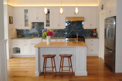 Mid-sized elegant l-shaped medium tone wood floor eat-in kitchen photo in San Francisco with a farmhouse sink, shaker cabinets, white cabinets, wood countertops, blue backsplash, subway tile backsplash, stainless steel appliances and an island