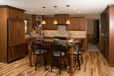 Example of a mid-sized classic l-shaped light wood floor open concept kitchen design in Minneapolis with an undermount sink, recessed-panel cabinets, medium tone wood cabinets, granite countertops, beige backsplash, porcelain backsplash and paneled appliances
