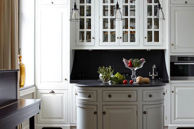 Пример оригинального дизайна: кухня в классическом стиле с белыми фасадами, черным фартуком, техникой из нержавеющей стали, островом, фасадами с выступающей филенкой и паркетным полом среднего тона