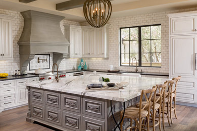 Inspiration for a mid-sized mediterranean l-shaped medium tone wood floor kitchen remodel in Orange County with a farmhouse sink, raised-panel cabinets, white cabinets, white backsplash, stainless steel appliances, an island, solid surface countertops and subway tile backsplash
