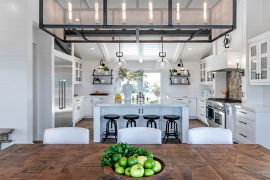 Diseño de cocina campestre de tamaño medio con armarios estilo shaker, puertas de armario blancas, encimera de mármol, salpicadero blanco, salpicadero de madera, electrodomésticos de acero inoxidable, suelo de madera en tonos medios, una isla, suelo marrón y encimeras blancas