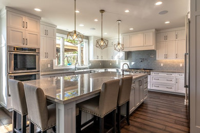 Huge trendy u-shaped dark wood floor and brown floor eat-in kitchen photo in Orange County with a farmhouse sink, shaker cabinets, white cabinets, quartzite countertops, gray backsplash, ceramic backsplash, stainless steel appliances, an island and beige countertops