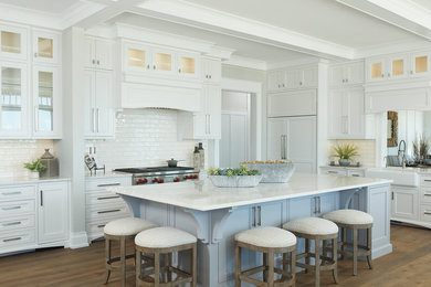 Example of a beach style l-shaped dark wood floor and brown floor kitchen design in Grand Rapids with a farmhouse sink, shaker cabinets, white cabinets, white backsplash, subway tile backsplash, stainless steel appliances, an island and white countertops