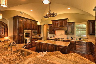 Inspiration for a large timeless u-shaped medium tone wood floor kitchen pantry remodel in Dallas with an undermount sink, raised-panel cabinets, dark wood cabinets, granite countertops, beige backsplash, stone tile backsplash, stainless steel appliances and two islands