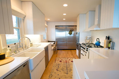 Example of a mid-sized beach style u-shaped medium tone wood floor eat-in kitchen design in Other with a farmhouse sink, flat-panel cabinets, white cabinets, white backsplash, subway tile backsplash, stainless steel appliances and solid surface countertops