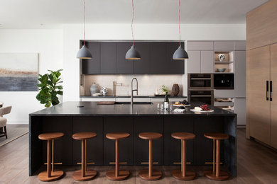 Example of a trendy l-shaped medium tone wood floor and brown floor eat-in kitchen design in Austin with an integrated sink, flat-panel cabinets, black cabinets, beige backsplash, stainless steel appliances, an island and black countertops