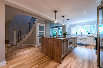 This is an example of a large contemporary l-shaped kitchen with a belfast sink, shaker cabinets, white cabinets, white splashback, stainless steel appliances, light hardwood flooring, an island, beige floors and brown worktops.