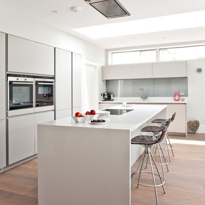 Inspiration for a contemporary l-shaped medium tone wood floor kitchen remodel in Surrey with an undermount sink, flat-panel cabinets, white cabinets, gray backsplash, glass sheet backsplash, paneled appliances and an island