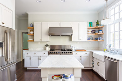 Photo of a large classic u-shaped kitchen pantry in Raleigh with a belfast sink, shaker cabinets, white cabinets, marble worktops, stainless steel appliances, dark hardwood flooring, an island, white splashback, marble splashback, brown floors and white worktops.