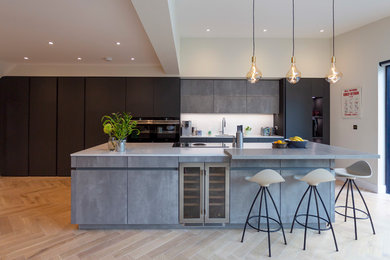 Photo of a large modern single-wall open plan kitchen in Surrey with an integrated sink, flat-panel cabinets, grey cabinets, quartz worktops, white splashback, black appliances, light hardwood flooring, an island, brown floors and white worktops.