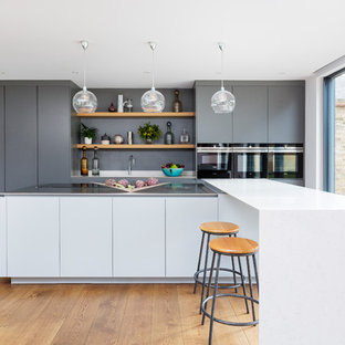 This is an example of a contemporary galley kitchen in London with flat-panel cabinets, grey cabinets, grey splashback, stainless steel appliances, medium hardwood flooring, an island, brown floors and white worktops.