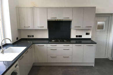 Photo of a modern u-shaped open plan kitchen in Oxfordshire with flat-panel cabinets, grey cabinets, composite countertops, black splashback, white appliances, porcelain flooring, grey floors and black worktops.