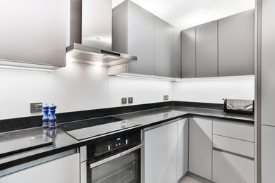 This is an example of a medium sized contemporary u-shaped kitchen in London with a submerged sink, flat-panel cabinets, grey cabinets, composite countertops, grey splashback, glass sheet splashback, stainless steel appliances, porcelain flooring, beige floors and black worktops.