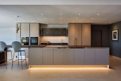 Large contemporary single-wall open plan kitchen in West Midlands with a built-in sink, flat-panel cabinets, grey cabinets, granite worktops, black appliances, limestone flooring, an island, beige floors and grey worktops.