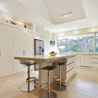 Photo of a contemporary u-shaped eat-in kitchen in Melbourne with flat-panel cabinets, white cabinets, window splashback, stainless steel appliances, with island, beige floor and beige benchtop.