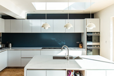 Inspiration for a medium sized contemporary l-shaped kitchen/diner in Surrey with a single-bowl sink, flat-panel cabinets, white cabinets, quartz worktops, blue splashback, glass sheet splashback, black appliances, an island and white worktops.