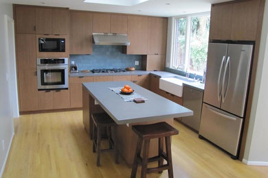 Example of a mid-sized trendy l-shaped light wood floor eat-in kitchen design in San Francisco with a farmhouse sink, flat-panel cabinets, light wood cabinets, solid surface countertops, gray backsplash, ceramic backsplash, stainless steel appliances and an island