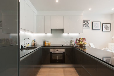 This is an example of a small contemporary u-shaped open plan kitchen in London with a double-bowl sink, flat-panel cabinets, black cabinets, glass sheet splashback, stainless steel appliances, light hardwood flooring, no island and beige floors.
