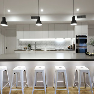 Photo of a contemporary l-shaped kitchen in Melbourne with an integrated sink, flat-panel cabinets, white cabinets, white splashback, stainless steel appliances, light hardwood floors, with island, beige floor and black benchtop.