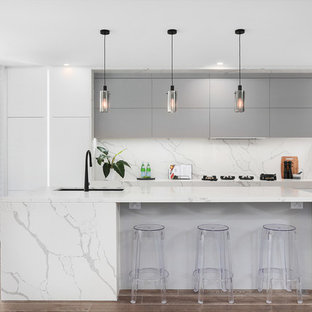 Mid-sized contemporary galley open plan kitchen in Sydney with a double-bowl sink, flat-panel cabinets, grey cabinets, marble benchtops, white splashback, marble splashback, black appliances, light hardwood floors, multiple islands, white benchtop and brown floor.