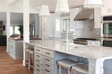 Example of a mid-sized transitional galley medium tone wood floor and brown floor kitchen design in St Louis with a farmhouse sink, recessed-panel cabinets, gray cabinets, gray backsplash, subway tile backsplash, stainless steel appliances and two islands