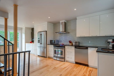 Example of a large trendy l-shaped light wood floor eat-in kitchen design in Montreal with flat-panel cabinets, white cabinets, granite countertops, white backsplash, stainless steel appliances, an island, black countertops, an undermount sink and glass tile backsplash