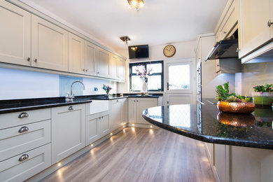 Small traditional u-shaped enclosed kitchen in Essex with a belfast sink, shaker cabinets, light wood cabinets, granite worktops, glass sheet splashback, black appliances, laminate floors, brown floors and multicoloured worktops.