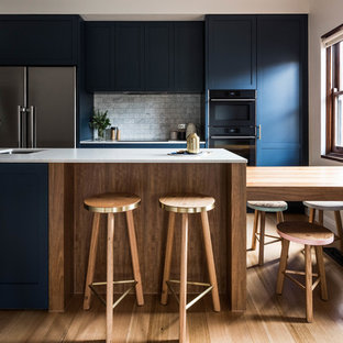 This is an example of a transitional galley kitchen in Melbourne with an undermount sink, shaker cabinets, blue cabinets, grey splashback, marble splashback, black appliances, medium hardwood floors, with island, brown floor and grey benchtop.