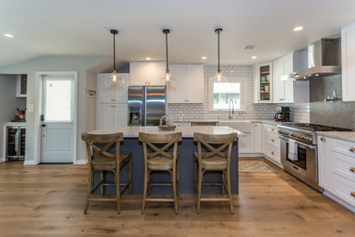 Large cottage l-shaped medium tone wood floor and brown floor eat-in kitchen photo in Los Angeles with a farmhouse sink, shaker cabinets, white cabinets, marble countertops, white backsplash, subway tile backsplash, stainless steel appliances, an island and gray countertops