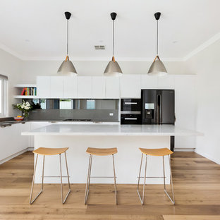 Design ideas for a contemporary l-shaped kitchen in Sydney with a drop-in sink, flat-panel cabinets, white cabinets, grey splashback, black appliances, light hardwood floors, with island and grey benchtop.