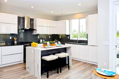 This is an example of a medium sized contemporary u-shaped kitchen/diner in Orange County with flat-panel cabinets, white cabinets, engineered stone countertops, black splashback, ceramic splashback, black appliances, light hardwood flooring, an island, brown floors, black worktops and a double-bowl sink.