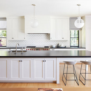 This is an example of a large classic galley open plan kitchen in San Francisco with a submerged sink, white cabinets, white splashback, metro tiled splashback, stainless steel appliances, light hardwood flooring, an island, shaker cabinets, beige floors, composite countertops and black worktops.
