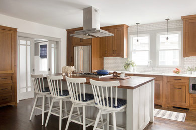 Example of a transitional l-shaped medium tone wood floor and brown floor kitchen design in Boston with an undermount sink, shaker cabinets, medium tone wood cabinets, wood countertops, white backsplash, subway tile backsplash, stainless steel appliances, an island and brown countertops