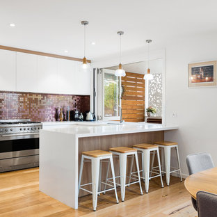 Photo of a mid-sized contemporary u-shaped eat-in kitchen in Sydney with an undermount sink, white cabinets, metallic splashback, mosaic tile splashback, stainless steel appliances, light hardwood floors, solid surface benchtops, with island, brown floor and white benchtop.