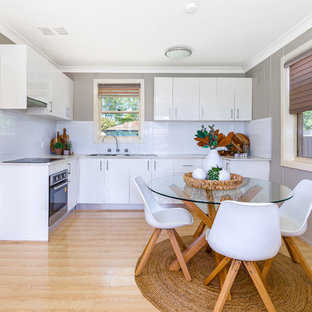 This is an example of a transitional l-shaped eat-in kitchen in Sydney with a double-bowl sink, flat-panel cabinets, white cabinets, white splashback, black appliances, light hardwood floors, beige floor and beige benchtop.