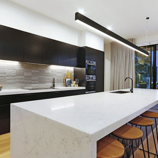 This is an example of a large contemporary galley open plan kitchen in Melbourne with an undermount sink, flat-panel cabinets, black cabinets, quartz benchtops, grey splashback, ceramic splashback, stainless steel appliances, light hardwood floors, with island, beige floor and white benchtop.
