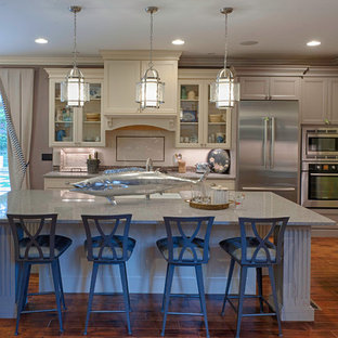 Photo of a mid-sized beach style l-shaped open plan kitchen in DC Metro with a farmhouse sink, recessed-panel cabinets, white cabinets, terrazzo benchtops, white splashback, porcelain splashback, stainless steel appliances, medium hardwood floors, with island and brown floor.
